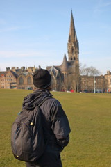 Turysta w ciemnym ubraniu, z plecakiem na plecach, tyłem, przygląda się wysokiej wieży zabytkowego kościoła w Edynburgu, Szkocja, zielony trawnik, zabudowa miejska w tle, błękitne niebo - obrazy, fototapety, plakaty