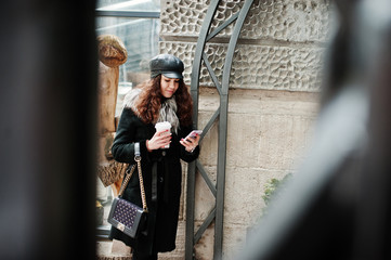 Curly mexican girl in leather cap and plastic cup of coffee at hand walking at streets of city.