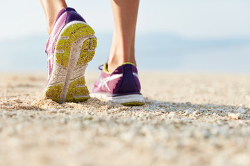 Cropped shot of female`s legs in purple training shoes stands at sandy beach on coastline, being in...