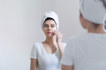 Young 25-year-old woman caring of her skin on the face, standing near mirror in the bathroom, uses a cotton disc and tones the face. Girl moisturizes the skin after a shower.