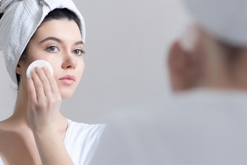 Young brunette woman with towel on hair removing makeup from her face, skin care concept.