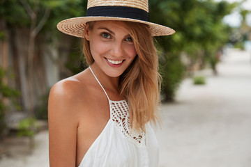 Horizontal shot of happy beautiful female with positive smile, wears summer hat and white dress, has walk outdoor, enjoys good rest during holidays and hot sunny weather. Vacation and lifestyle