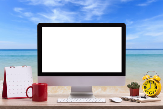 Modern desktop computer, Coffee cup, alarm clock, notebook and calendar on wooden table and view of tropical beach. Saved with clipping path. Work and holiday concept