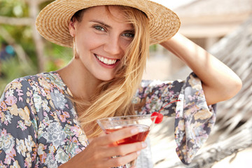 Beautiful happy young woman in summer style, has appealing appearance, smiles broadly, drinks fresh red cherry coctail, enjoys recreation after hard work, has trip abroad in hot exotic place