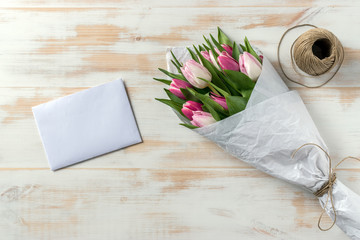 Bouquet of pink tulips wrapped in white paper and white envelope on a wooden table. Flat lay. Love concept.