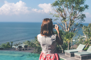 Fototapeta na wymiar Young woman taking photos on the cliff with a beautiful ocean background at sunny day. Bali island.