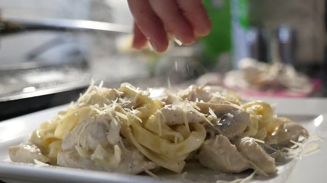 An impressive closeup of a tasty dish cooked from makaroni, meat slices and cheese chips. Female hands sprinkle it and takes to an oven 