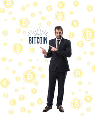bearded smiling businessman in suit pointing at word bitcoin isolated on white