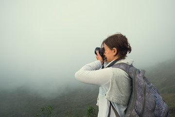 Caucasian woman making photo standing on the top of mountain. It is very cloudy but she tries to shoot.