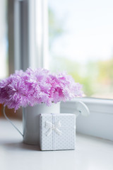 Small white watering can with tender bouquet of beautiful pink carnation and gift box near window in daylight