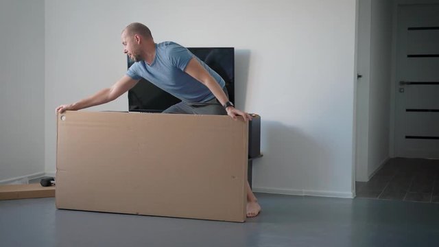 adult man is turning and lying on a floor big cardboard box in an room of apartment, cutting packing tape
