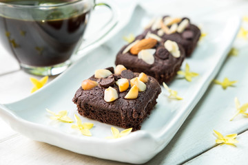 Brownie with macadamia and chocolate ship and almond on top with yellow petal flower on white plate on old white wooden table , there is cup of hot coffee in the background