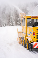 Big yellow snowplough with screw and helix is cleaning the road while a powerful stream of white snow go out