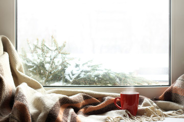 cozy home atmosphere in the winter/ red mug of hot drink on a windowsill with a warm blanket, when...