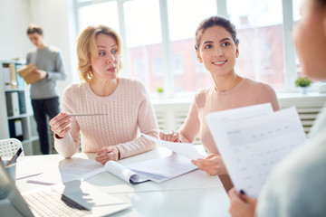 Young manager listening attentively to colleague while discussing papers by workplace