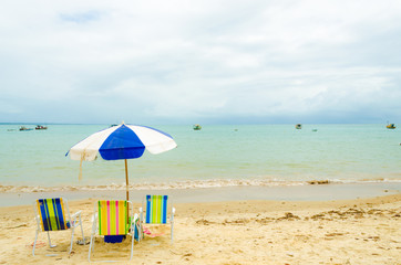 Isolated beach with three colorful beach chairs and a blue and white umbrella. A small wave breaks in the sand and some boats are in the sea. Cloudy sky.