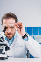 scientist in white coat and eyeglasses looking through microscope on reagent in laboratory