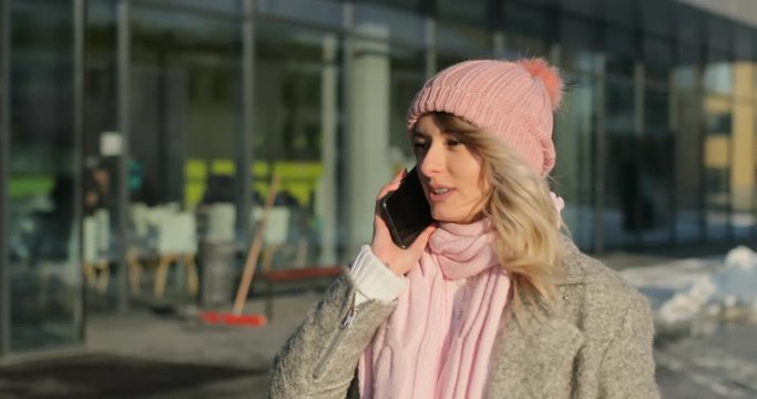 Winter time in big city, of charming girl answering call, talking on the smart phone, walking on street. Enjoying snowfall, woman wearing a winter coat and pink scarf and hat, new year mood