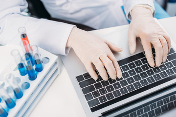 partial view of scientist in lab coat and medical gloves typing on laptop at workplace with reagents