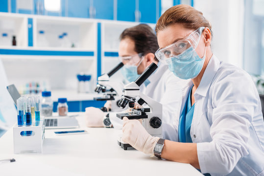selective focus of scientists in medical masks and goggles looking through microscopes on regents in lab