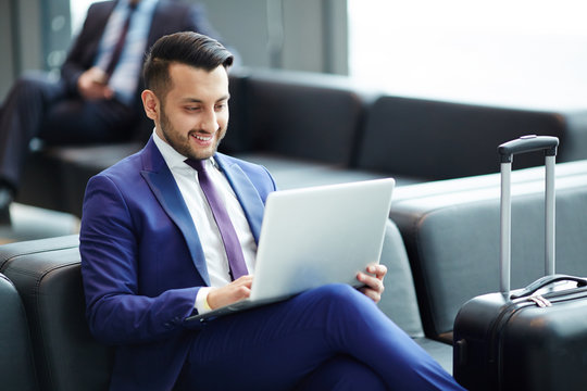 Elegant young man with laptop looking at its display with smile while networking in lounge