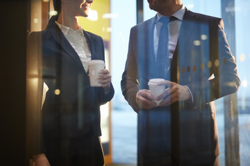 Two colleagues in formalwear having coffee and talking behind transparent wall inside modern building
