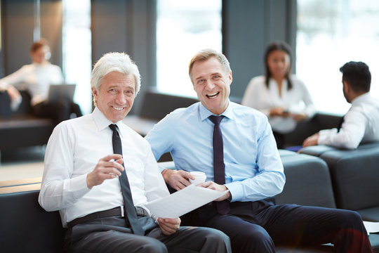 Two laughing mature businessmen looking at camera while having meeting in airport lounge