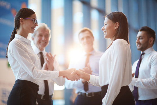 Two Young Intercultural Businesswomen Shaking Hands And Looking At One Another With Three Men Applauding Near By