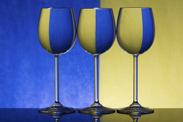 Three wineglasses on a shiny surface with water that distort yellow and blue background