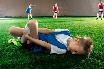 Little player lying on pitch and suffering from pain in his knee or leg after falling