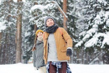 Fototapeta na wymiar Young couple in winterwear walking in snowdrift and enjoying time in the forest on winter weekend