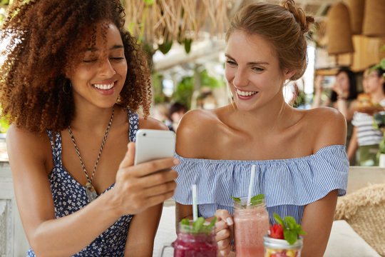 Dark skinned curly woman with positive expression shows photos to her best friend on smart phone, drink smoothie. Lesbian couple recreat in restaurant with modern gadget. Friendship concept.
