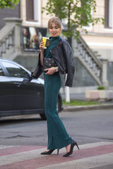 Fototapeta premium Beautiful rich luxury looking blonde caucasian business woman in casual green outfit standing on a european streets, holding coffee and purse. Daylight, outdoors.