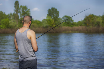 Young handsome brutal caucasian man in casual outfit fishing on a lake as a hobby, trying to catch carp on spinning rod alone. Summer weather, sunset, natural landscapes. HDR toned image