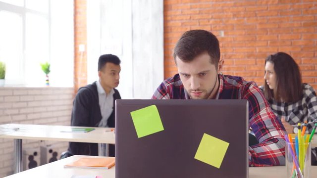 young attractive man sitting in front of computer and typing on laptop. The concentrated man looks closely at the monitor. Shooting in the office. in the background, the extras from the actors. 4k