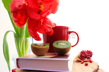 Morning concept. Healthy lifestyle. Red flat lay: fresh fruits kiwi, pomegranate. Red tulips and cup on the book. Close up