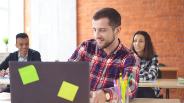 young attractive man sits in front of a computer and communicates with colleagues. a friendly atmashere in the classroom. people are laughing. Shooting in the office. in the background, the extras