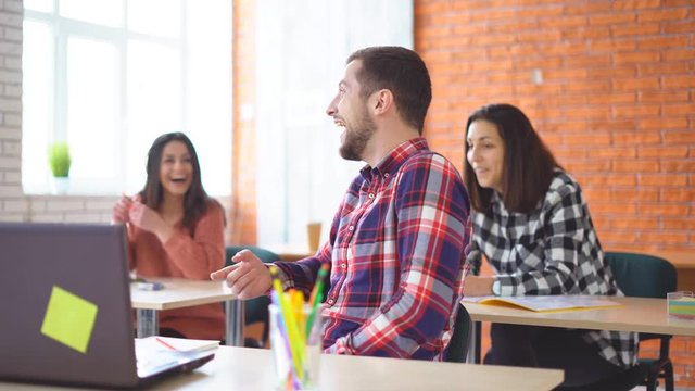 young attractive man sits in front of a computer and communicates with colleagues. a friendly atmashere in the classroom. people are laughing. Shooting in the office. in the background, the extras