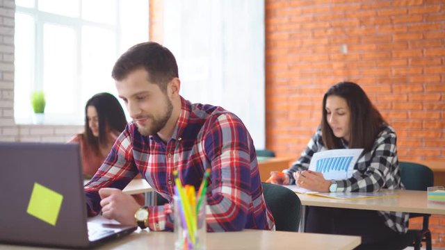 young attractive man sits in front of a computer and communicates with colleagues. a friendly atmashere in the classroom. people are laughing. Shooting in the office. in the background, the extras