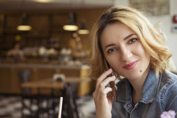 Blonde (with dark roots) caucasian woman in casual summer outfit at the cafe. Grey dress and jeans jacket. Woman got natural day makeup and curly hairstyle. She talks on her phone, working, make calls