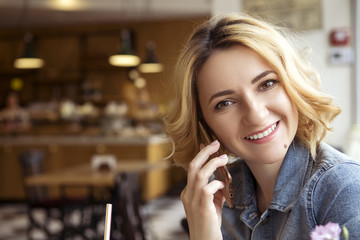 Blonde (with dark roots) caucasian woman in casual summer outfit at the cafe. Grey dress and jeans jacket. Woman got natural day makeup and curly hairstyle. She talks on her phone, working, make calls