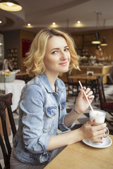 Blonde (with dark roots) caucasian woman in casual summer outfit at the cafe. Grey dress and jeans jacket. Woman got natural day makeup and curly hairstyle. She drinks coffee latte and eats croissant
