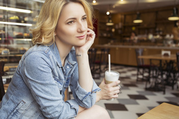 Blonde (with dark roots) caucasian woman in casual summer outfit at the cafe. Grey dress and jeans jacket. Woman got natural day makeup and curly hairstyle. She drinks coffee latte and eats croissant