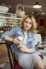 Blonde (with dark roots) caucasian woman in casual summer outfit at the cafe. Grey dress and jeans jacket. Woman got natural day makeup and curly hairstyle. She drinks coffee latte and eats croissant
