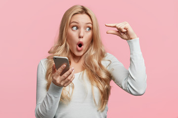 Shocked European female with blonde hair, shows something very tiny, holds smart phone in hands,...