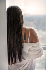 portrait of a girl with long hair in a white shirt standing back to us in front of a large window. Woman with tattoo on shoulder  © Viktoria