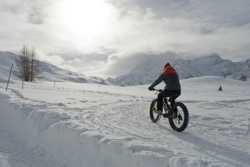 Fototapeta premium man use electric bicycle, e-bike, ebike, pedal on snow covered road, downhill mountain, specific bike with wide wheels to go on snow, called fatbike, winter, cold, alps, Simplon Pass, Switzerland