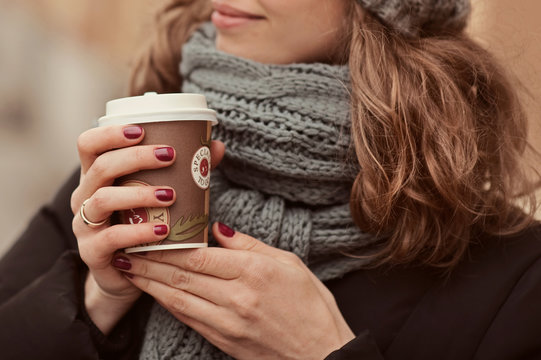 Young girl in warm scarf on the street holding coffee to go or take away while walking in the park, mock-up of coffee cup