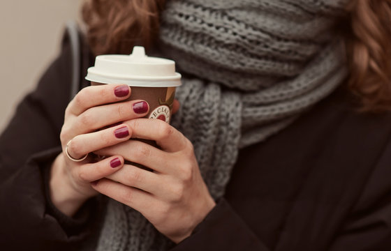 Young girl in warm scarf on the street holding coffee to go or take away while walking in the park, mock-up of coffee cup