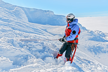 Fototapeta na wymiar Equipped climber ascent by snowy slope with climbing rope on the top of peak in snowy alpine mountains. Life guard professional man on the work in high mountains. Action in hard conditions scene.